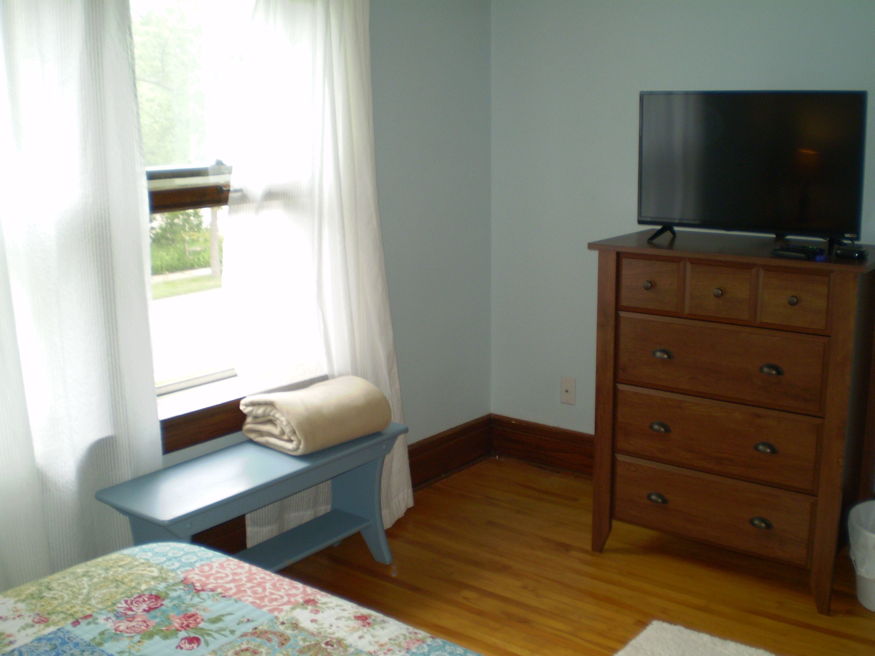 Une chambre à coucher avec un lit, une commode en bois, un téléviseur posé dessus, une fenêtre avec des rideaux et un banc près de la fenêtre.