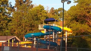 A playground with multiple slides and a covered play area, surrounded by trees and a fence.