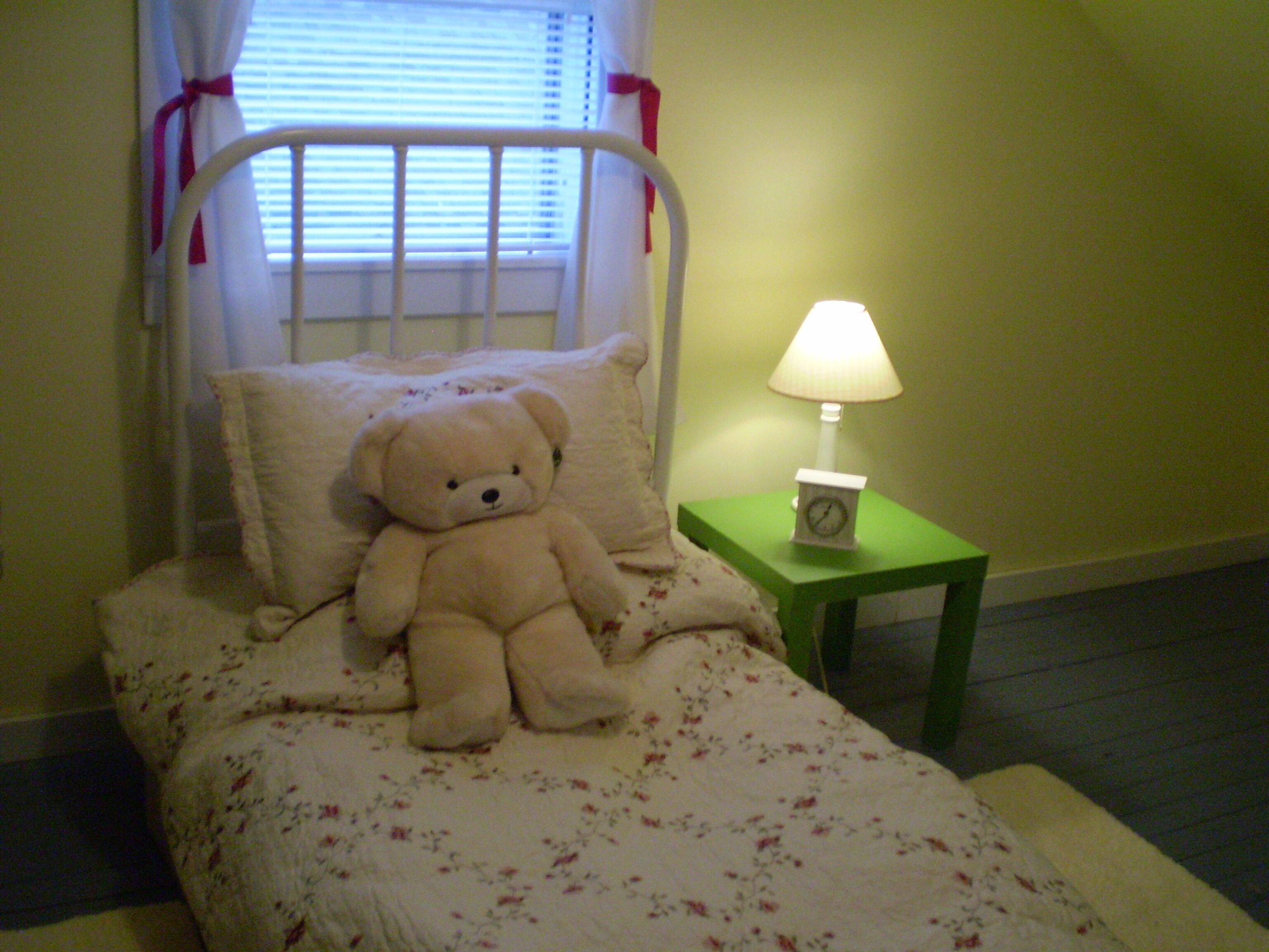 A bed with a floral bedspread, a white metal headboard, a teddy bear, a bedside lamp, and a clock on a green nightstand.
