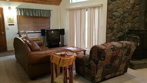 A cozy living room with a brown leather sofa, a wooden coffee table, and a patterned armchair. A television is on a stand, and there is a stone wall behind the armchair. - Pinecrestarearentals #8 Getaway Fall/labor day 3rd night free just Book 2nights. (Cold Springs)