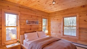 Un dormitorio de cabaña de madera con una cama, mesitas de noche y ventanas grandes.