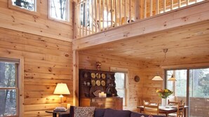 Una cabaña de madera espaciosa con un dormitorio en el altillo, una sala de estar acogedora con sofá y mesa de comedor, y grandes ventanales que ofrecen vistas del bosque circundante.