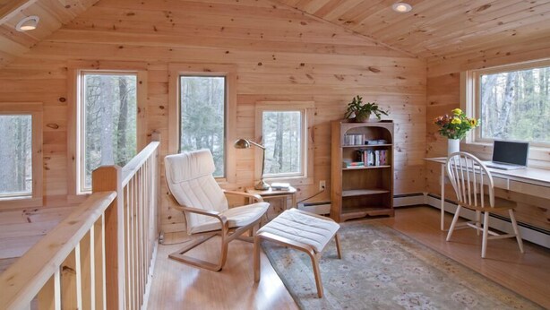 Interior acogedor de una cabaña con paredes y suelos de madera, un escritorio con una computadora portátil, una estantería y una zona de estar cómoda con sillas y un banco.