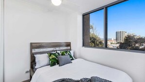 A bedroom with a large window, a bed with white bedding, and a view of buildings and trees.
