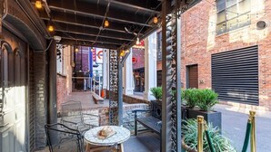 Un patio al aire libre con sillas y mesa de metal, rodeado de muros de ladrillo y una zona cubierta con luces colgantes.