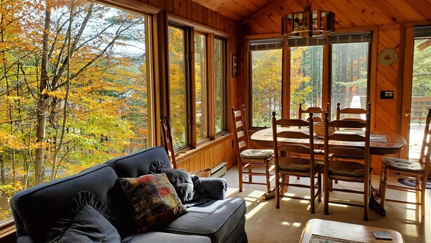 Interior acolhedor de uma cabana com teto de madeira, grandes janelas com vista para árvores de outono, um sofá com almofadas e uma mesa de jantar com cadeiras.