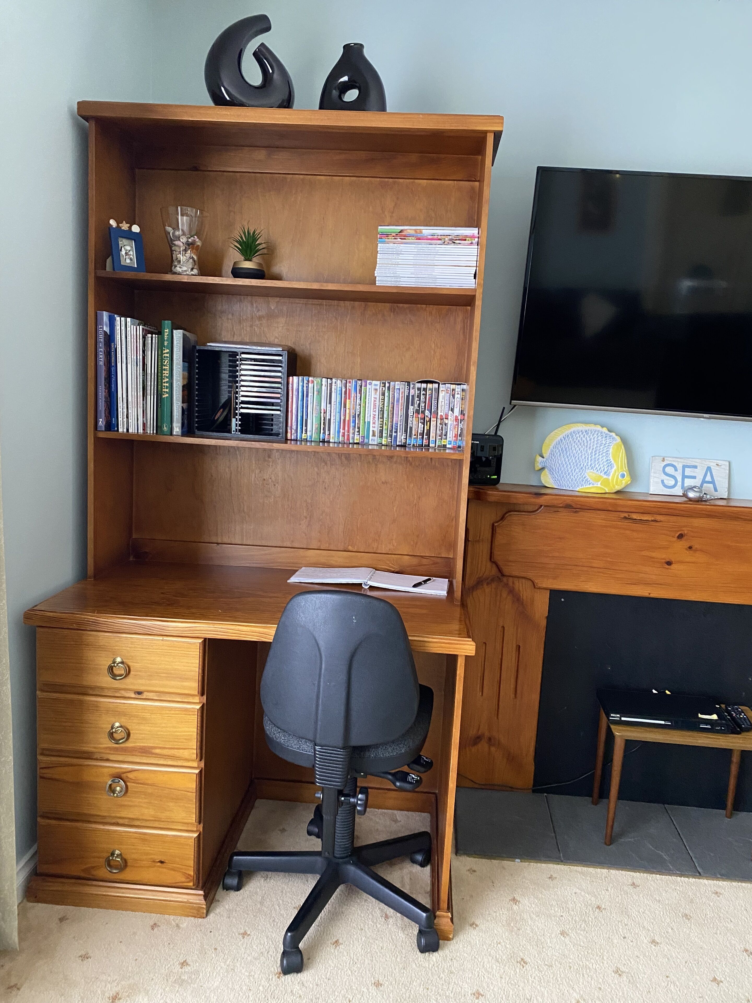 A wooden desk with a black office chair, a bookshelf with books and decorative items, a television, and a small shelf with a fish decoration and a sign that says "SEA".