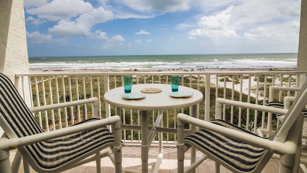 Un balcon avec deux chaises et une table, donnant sur une plage où l’on peut voir des vagues et un ciel nuageux.