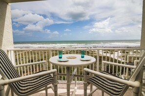 A balcony with two chairs and a table, overlooking a beach with waves and a cloudy sky.