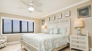 Une chambre à coucher avec un lit, des tables de chevet, un ventilateur de plafond et une grande fenêtre.