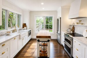 Una cocina con armarios blancos, electrodomésticos de acero inoxidable, una isla de madera y ventanas amplias con vistas a zonas verdes.
