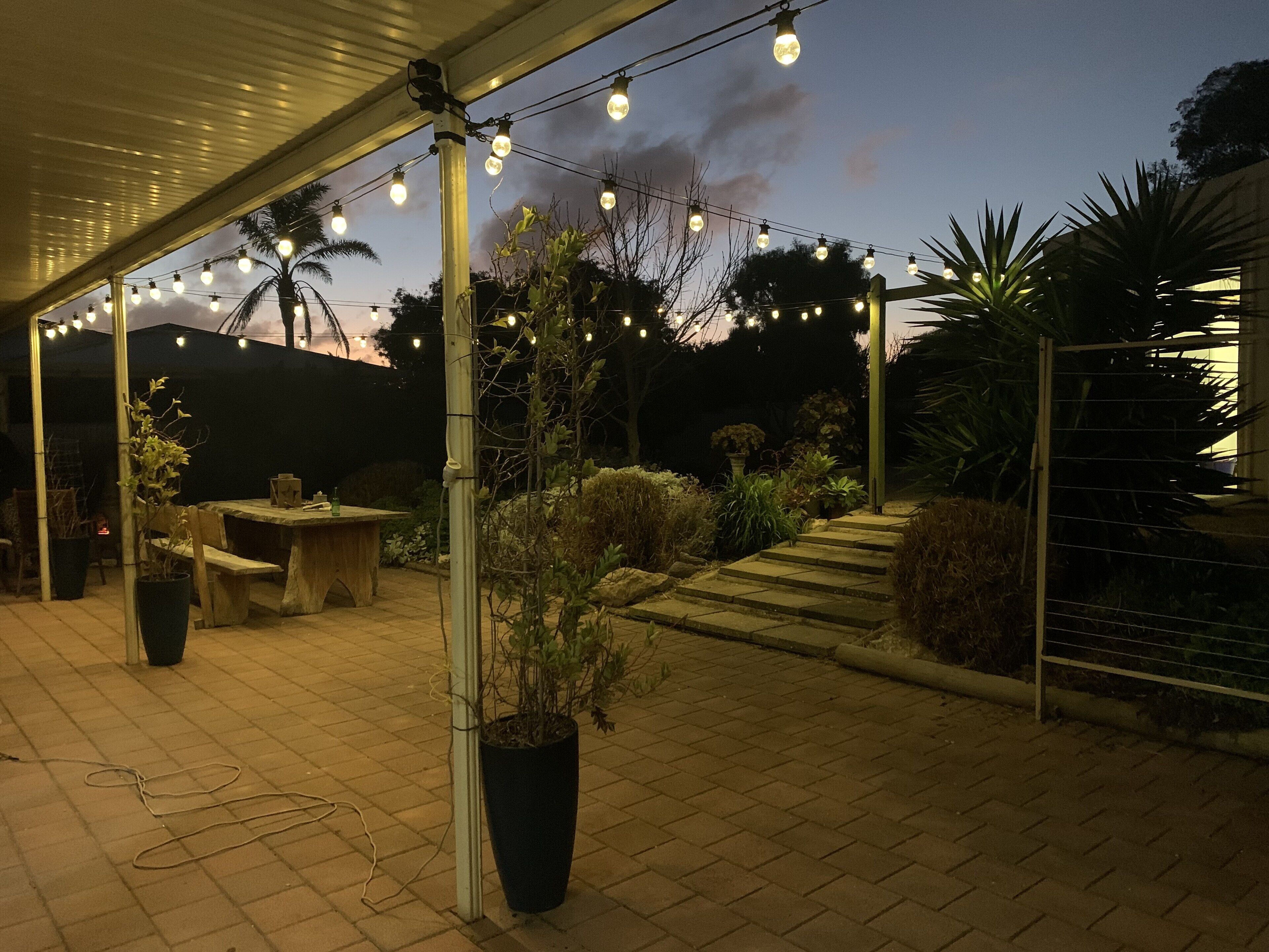 Un espace extérieur aménagé en terrasse, agrémenté de guirlandes lumineuses, de plantes en pot et d’une table en pierre.