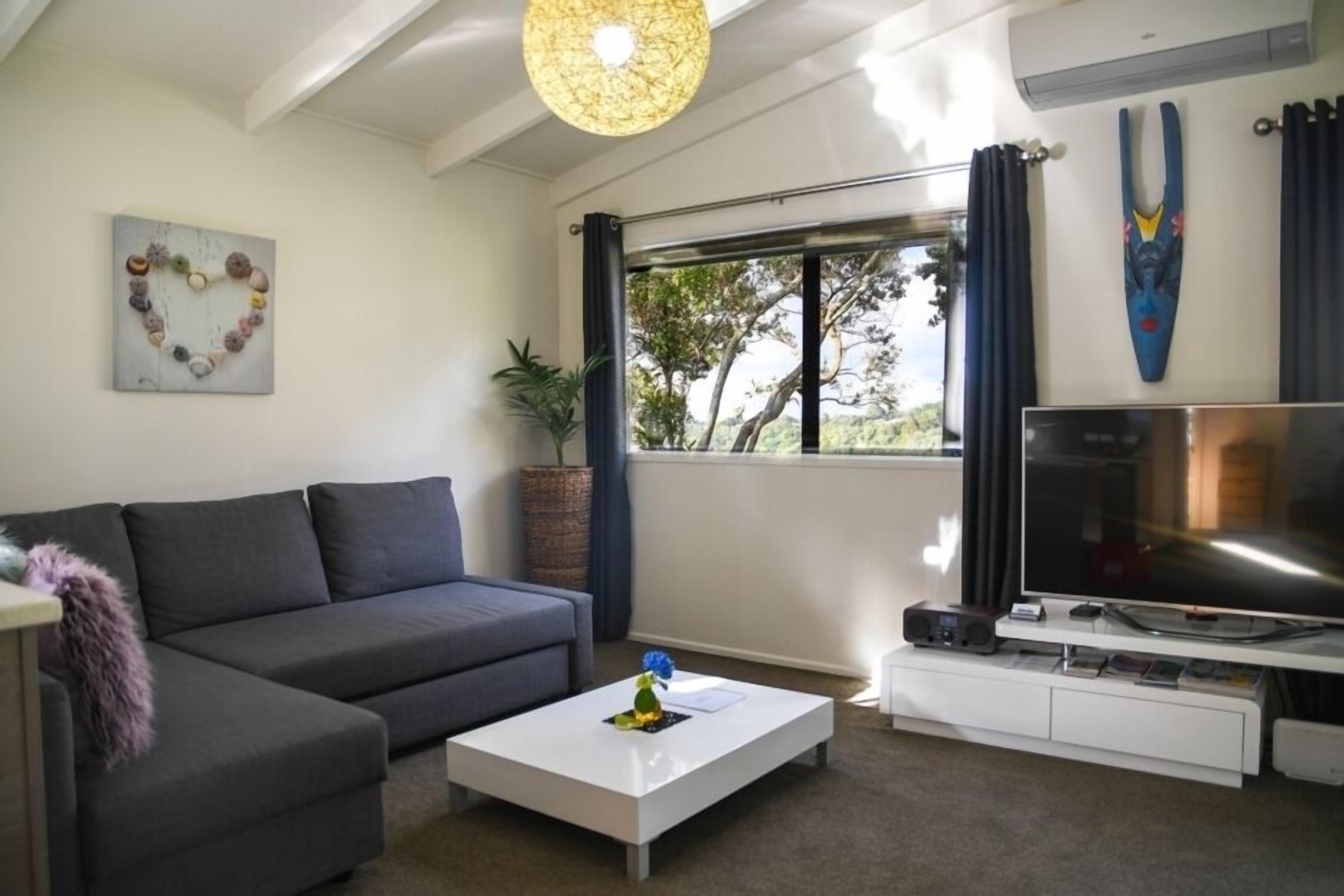 A modern living room with a grey sofa, a white coffee table, a flat-screen TV, and a large window with curtains.