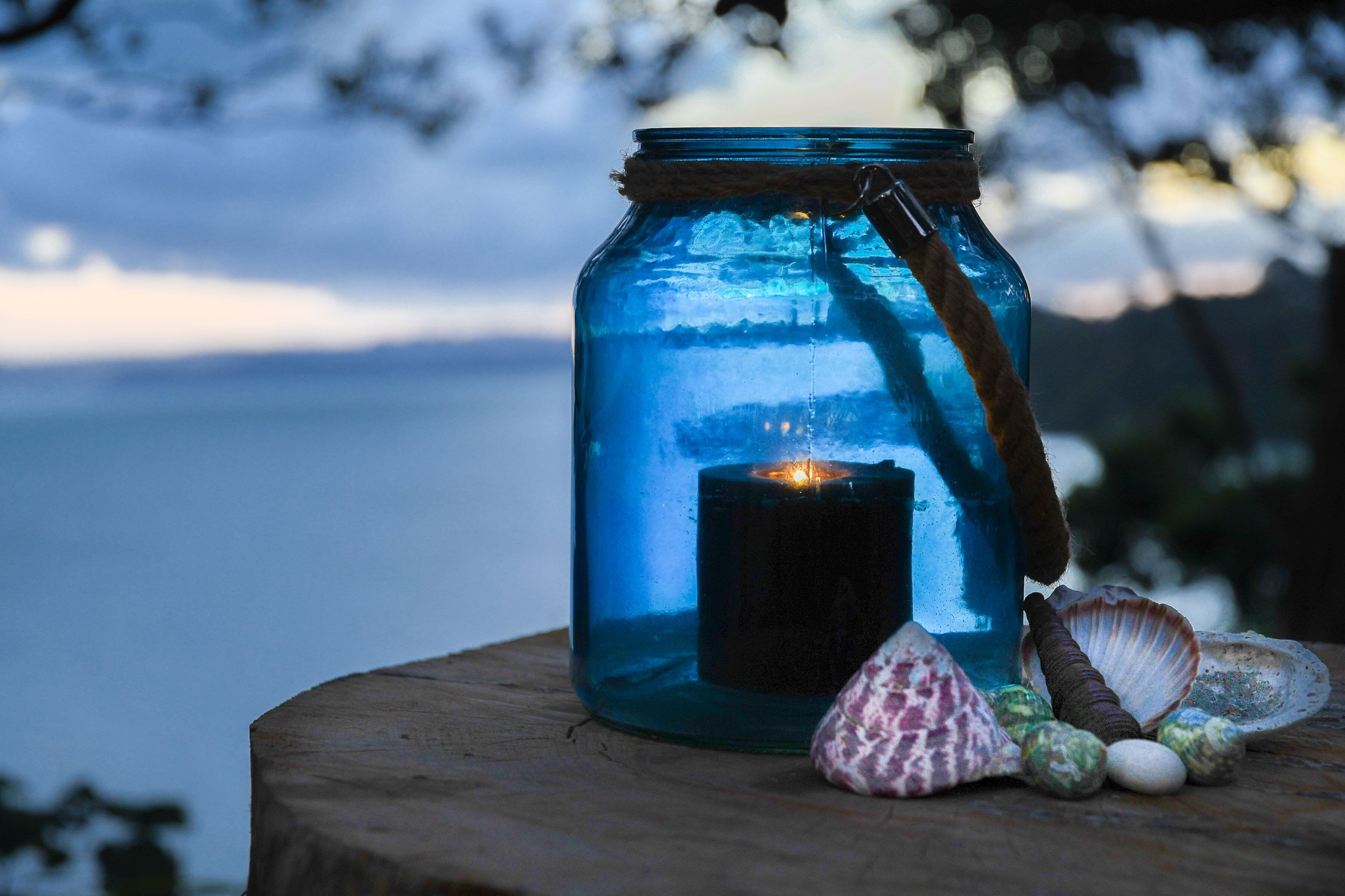 A blue glass jar with a candle inside, placed on a wooden surface with seashells around it.