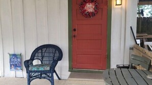 A red door with a wreath, a black wicker chair, and a wooden bench outside a building with a white exterior.