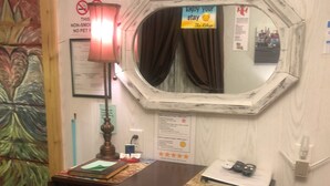 A wooden desk with a mirror, a lamp, and a small stool. There are papers and a remote control on the desk.