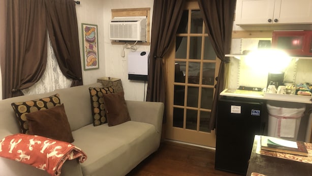A living room with a sofa, pillows, a blanket, a small kitchen area with a refrigerator, and a window with curtains.