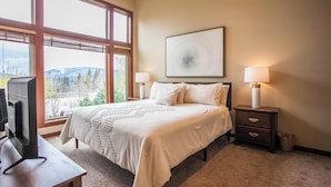Une chambre avec une grande fenêtre, un lit, une table de chevet, une lampe et un téléviseur.