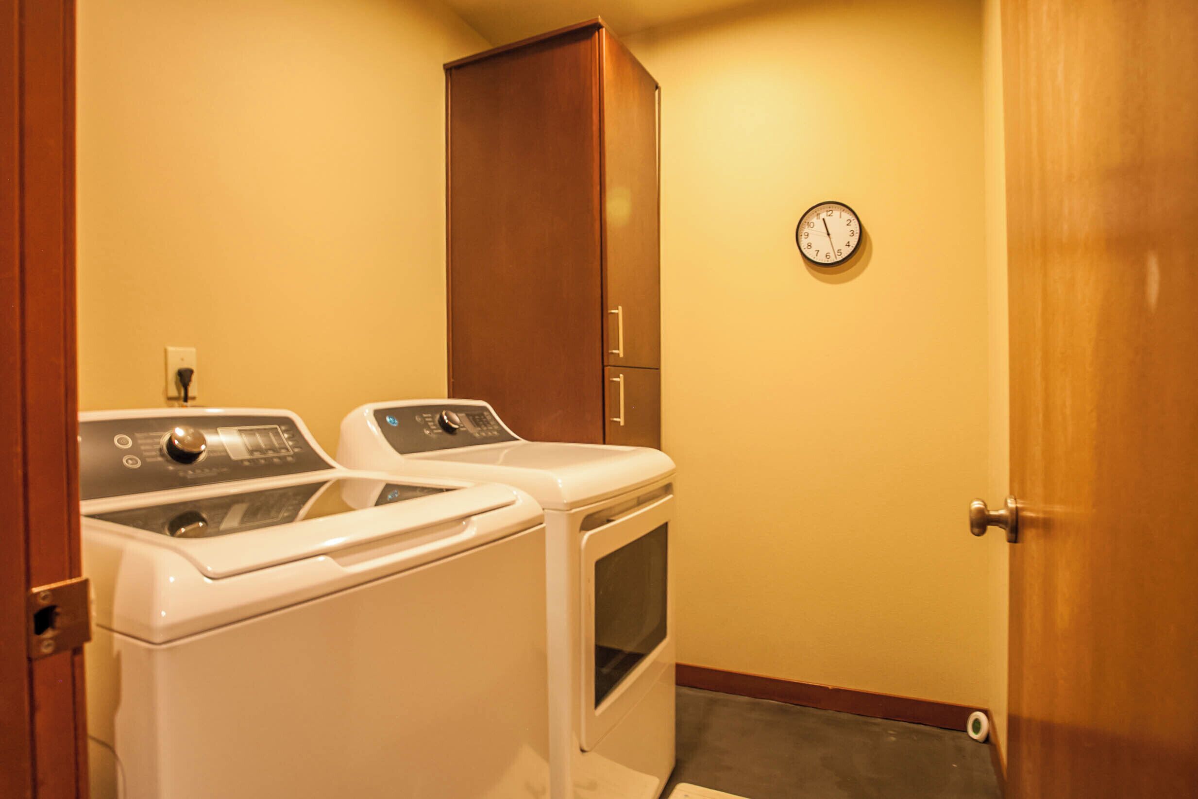 A laundry room with two washers and a dryer, a wooden cabinet, and a wall clock.
