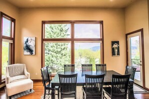A dining area with a table and chairs, large windows with a view of trees and mountains, and artwork on the walls.