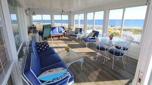 A covered outdoor seating area with blue and white furniture, a glass table with a vase of yellow flowers, and a view of the ocean. - 93 Phillips Rd SD246 (Sagamore Beach)
