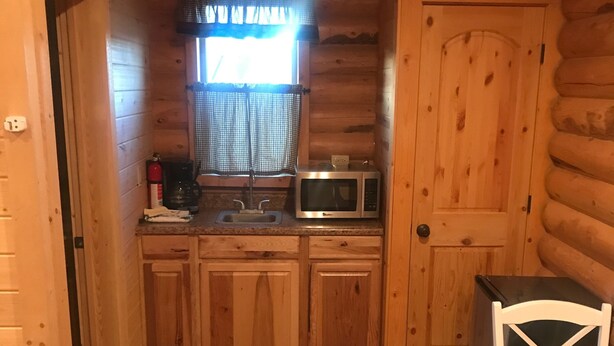 A compact kitchen with wooden cabinets, a microwave, a coffee maker, and a window with curtains. - Cabin 2 (Garden City)