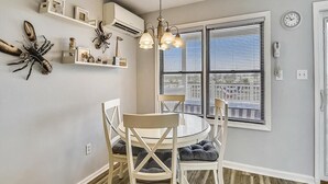 Un coin repas avec une table ronde, des chaises blanches et un lustre. On y trouve des étagères murales ornées d’objets décoratifs et une fenêtre avec des stores.