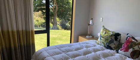 Un dormitorio con una cama grande, una mesita de noche con una lámpara y vistas a un jardín por la ventana.