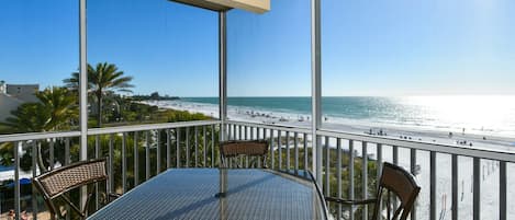 A balcony with a glass table and chairs, overlooking a beach with people and palm trees.