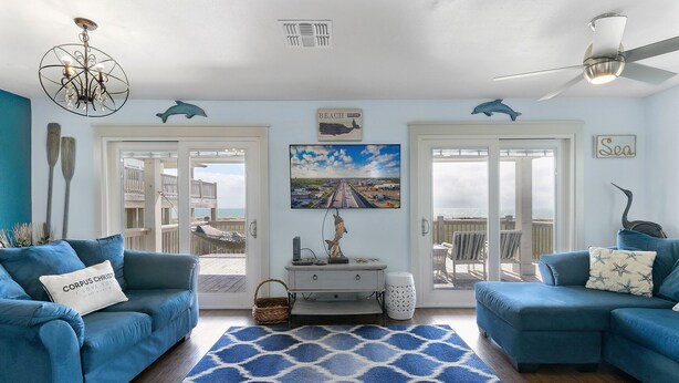 Een woonkamer met twee blauwe banken, een tapis plein, een televisie en een balkon met uitzicht op het strand.