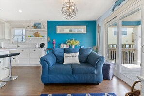 A modern living room with a blue sofa, a chandelier, and a view of the outside deck. - Reef on the Beach (Corpus Christi)