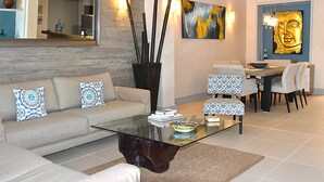 A modern living room with a glass coffee table, a sofa with patterned cushions, a dining area with patterned chairs, and a Buddha statue on the wall.