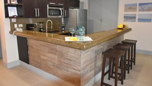 A modern kitchen with a granite countertop, stainless steel appliances, and wooden cabinetry. There are bar stools and a small island with a sink.