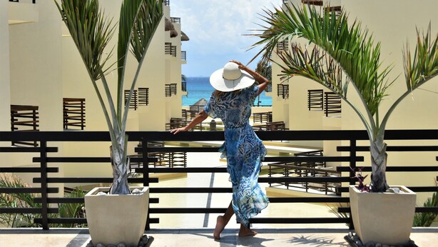 A woman in a blue dress and white hat walking on a balcony with a view of the ocean.