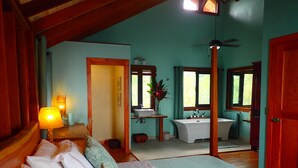 A bedroom with a bed, a bedside table, a lamp, a window, and a bathroom with a sink and bathtub.
