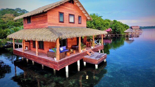A wooden house with a thatched roof on stilts over water, surrounded by lush greenery.