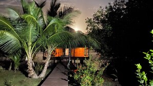 A pathway leading to a building with a thatched roof, surrounded by lush greenery and palm trees.