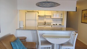 Una cocina compacta con refrigerador, microondas, estufa y lavamanos. Hay un pequeño comedor con una mesa y sillas blancas. Un letrero sobre la cocina dice "BEACHHOUSE".