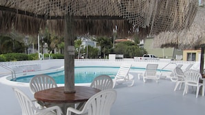 Un área junto a la piscina con sillas de plástico blancas y una mesa de madera, rodeada por un techo de paja.