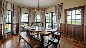 A dining room with a wooden table set for four, large windows with patterned curtains, and a chandelier.