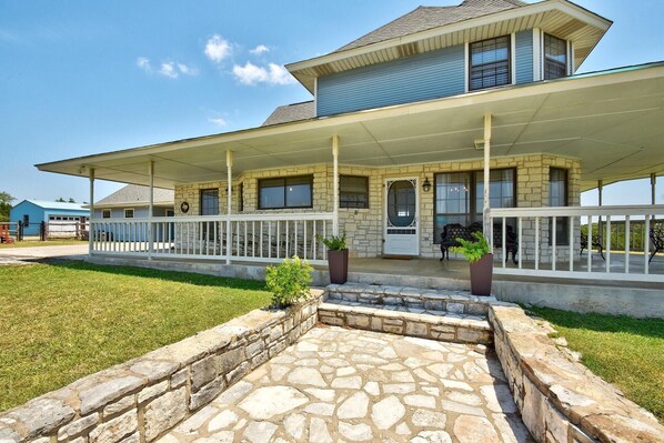 A two-story house with a stone foundation, a covered porch with white railings, and a stone patio in front. - Prosit Ranch - 300 Acre Private Ranch w/360 Degree Views & Wrap-Around Porch (Wimberley)