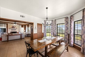 A spacious kitchen with a wooden dining table, a built-in island, and a large window with patterned curtains. - Prosit Ranch - 300 Acre Private Ranch w/360 Degree Views & Wrap-Around Porch (Wimberley)