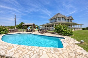 A swimming pool with stone tiles, a covered seating area, and a large house with a porch. - Prosit Ranch - 300 Acre Private Ranch w/360 Degree Views & Wrap-Around Porch (Wimberley)