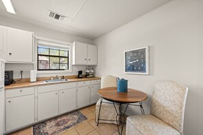 A kitchen with white cabinets, a sink, a window, a clock, and a small round table with a chair.