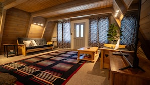A cozy attic bedroom with a wooden ceiling, a bed with a patterned bedspread, a desk with a computer, and a large window with curtains.