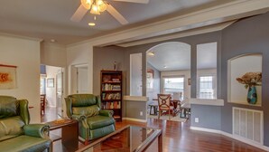 A spacious living room with a wooden floor, green leather sofas, a glass coffee table, and a bookshelf.