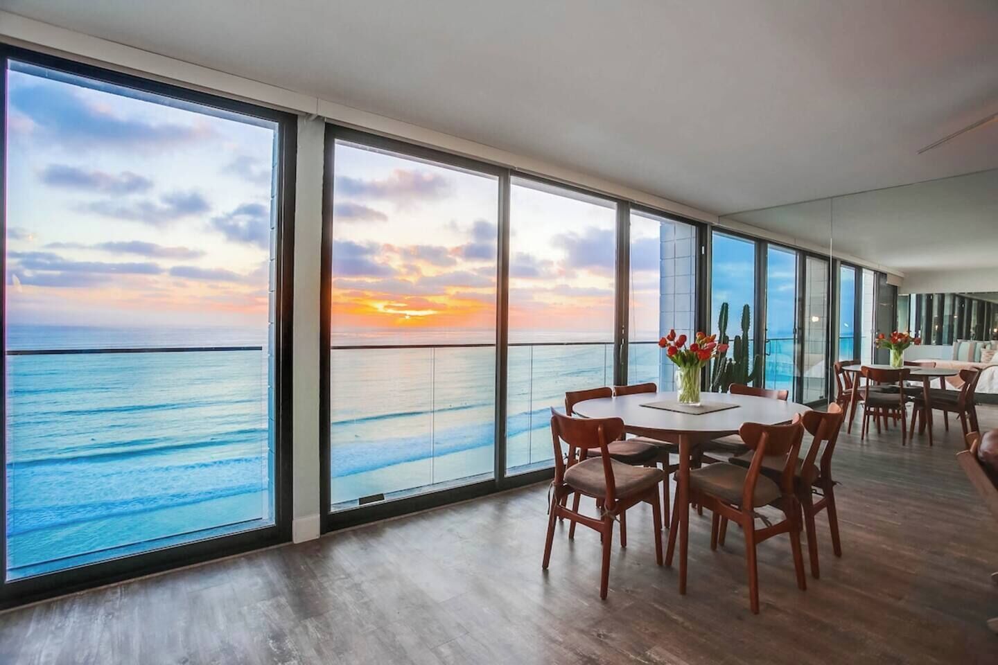 A room with a large window overlooking the sea, a round dining table with chairs, and a small table with a vase of flowers.