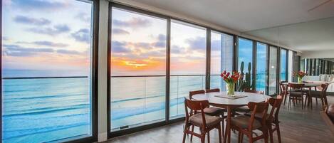 A room with a large window overlooking the sea, a round dining table with chairs, and a small table with a vase of flowers.
