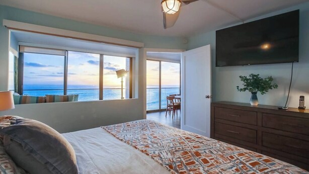 A bedroom with a large bed, a ceiling fan, a TV, a window with a view of the ocean, and a door leading to a balcony.