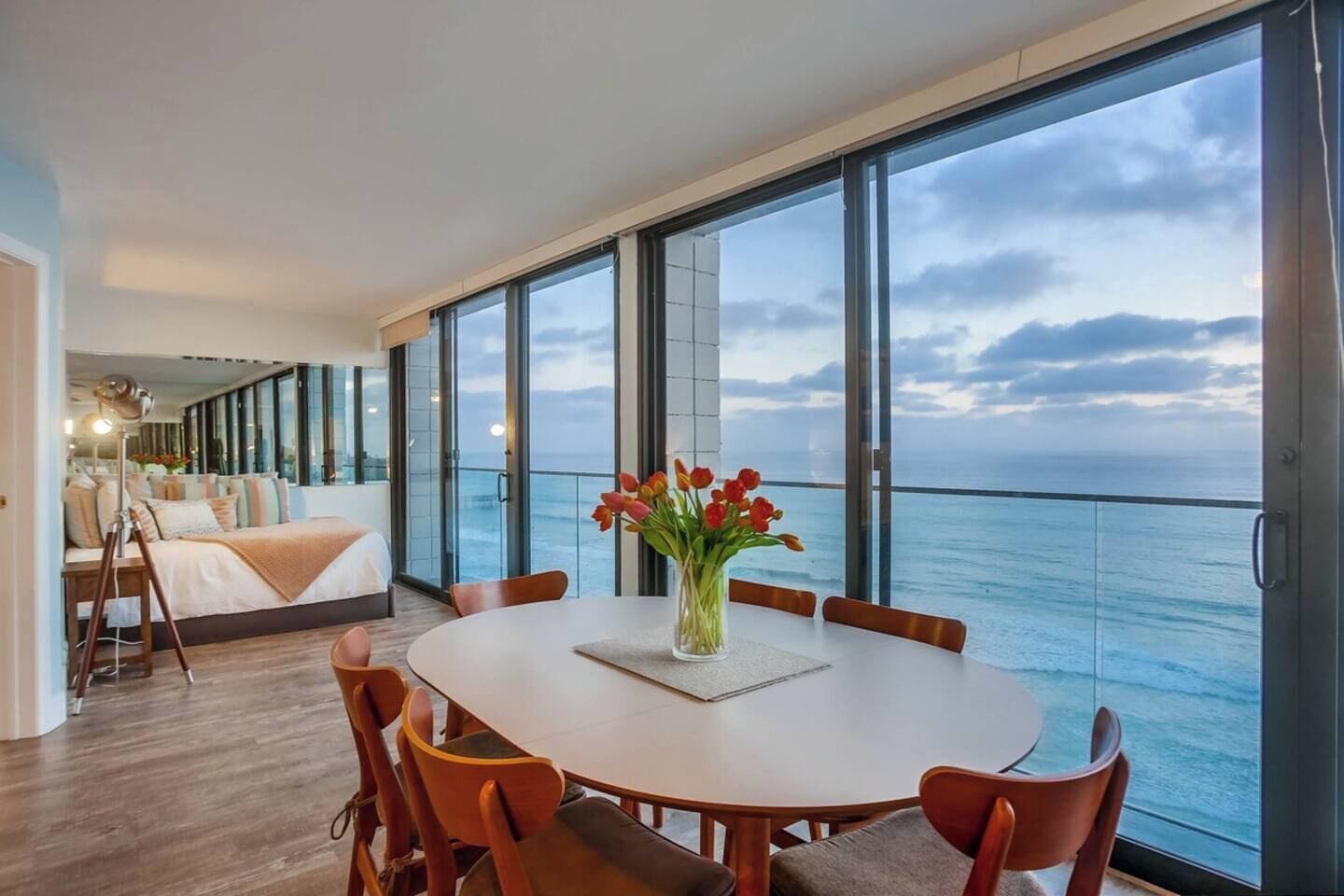 A modern room with a large window overlooking the sea, a round dining table with chairs, and a bed with a view.